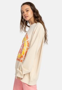 Beige oversized sweatshirt featuring colorful floral and text graphics in orange and yellow. Soft texture, long sleeves, ribbed cuffs.