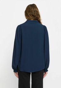 Blouse marine à manches longues, col montant boutonné, coutures d'épaules froncées et poignets élastiqués. Texture lisse, coupe légèrement ample.