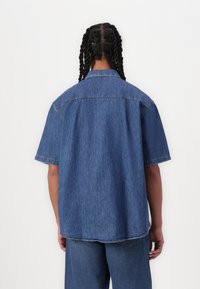 Cămașă din denim cu mâneci scurte, de culoare albastră, cu o croială relaxată, având guler, cusături vizibile și un tiv ușor curbat în partea de jos.