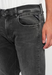 Black denim jeans with a slightly faded texture, five-pocket design, front button closure, and logo tag on the back pocket.