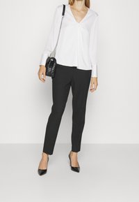 Bílá blůza s dlouhým rukávem a výstřihem do V, zdobená nařasením, v kombinaci s černými saka kalhotami a černými lodičkami s špičatou špičkou, držící černou kabelku.