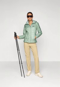 Φωτεινό πράσινο μονωμένο μπουφάν με κουκούλα, φερμουάρ εμπρός και διακριτικό καπιτονέ σχέδιο· μπεζ παντελόνι· μαύρα μπαστούνια πεζοπορίας· μπεζ αθλητικά παπούτσια· γυαλιά ηλίου.