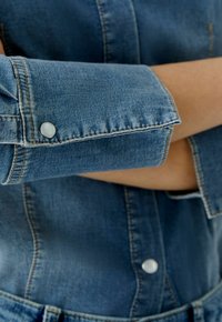 Denim shirt met mouwen in medium blauw met zichtbare stiksels. Manchet is opgerold met een zilveren drukknoopdetail. Soepele textuur met een lichte vervaging.