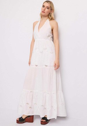 Donna che indossa un vestito lungo bianco senza maniche con dettagli in pizzo, in piedi contro uno sfondo chiaro e semplice, abbinato a sandali platform neri.