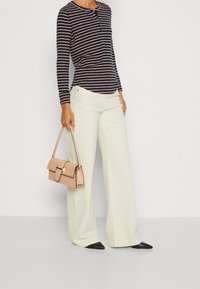 Pasiasta bluzka z długim rękawem w kolorze granatowym i beżowym, zestawiona z jasnoróżowymi szerokimi spodniami. Trzyma małą, usztywnioną torebkę w jasnoróżowym kolorze ze złotymi okuciami.