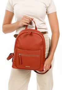 Femme tenant un petit sac à dos en cuir marron texturé avec une poche avant à fermeture éclair, portant une chemise blanche et un pantalon beige.
