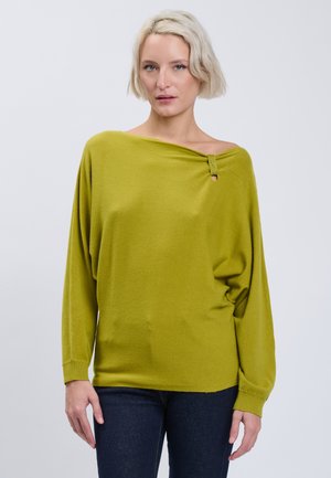 Frau mit kurzen blonden Haaren, die einen olivgrünen schulterfreien Pullover und dunkle Jeans trägt, vor einem schlichten Hintergrund stehend.