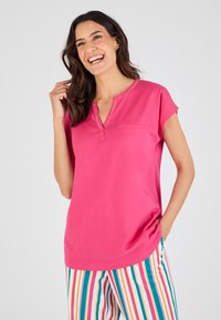 Haut rose à manches courtes avec un décolleté en trou de serrure et une seule poche sur la poitrine, fabriqué dans un tissu lisse. Associé à un pantalon rayé multicolore.