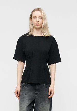 Ξανθιά γυναίκα που φοράει εφαρμοστό μαύρο τοπ με κοντά μανίκια και ξεθωριασμένο μαύρο τζιν, στέκεται μπροστά σε απλό ανοιχτό γκρι φόντο.