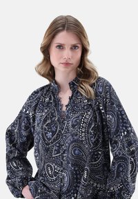 Una donna bionda con capelli mossi indossa una blusa a maniche lunghe di colore blu navy, decorata con motivi paisley e floreali bianchi e blu, con le mani sui fianchi.
