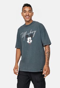 Camiseta gris de gran tamaño hecha de algodón, con un logo blanco de "Mickey" y un gráfico de Mickey Mouse en la parte frontal. Mangas cortas y cuello redondo.
