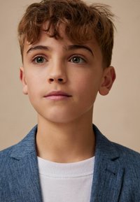 Blazer blu a trama sopra una camicia bianca a coste. In primo piano, un bambino con capelli castani ondulati e occhi verdi su uno sfondo chiaro.