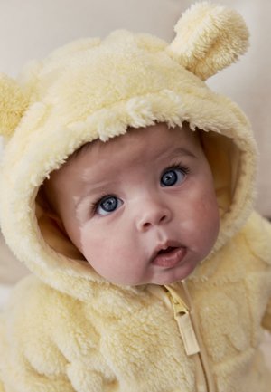 Bebé con ojos azules vestido con un suave conjunto amarillo con capucha y orejas de oso, mirando hacia arriba con expresión curiosa.