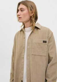 Beige corduroy overhemd met een knoopsluiting aan de voorkant, lange mouwen, een enkele borstzak en labeldetail nabij de zak. Gelaagd ribstof.