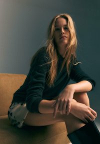 Un pull noir, un short à motifs et des bottes noires jusqu'aux genoux. Assise sur un canapé beige, avec de longs cheveux et une posture décontractée. Éclairage doux.