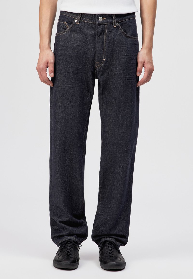 Homme portant un jean droit bleu foncé, un t-shirt blanc et des baskets noires, debout devant un fond clair uni.