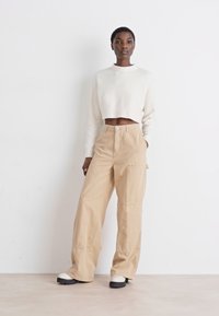 Pantalons larges beiges à taille haute avec des détails de couture visibles, associés à un pull cropped blanc. Les chaussures sont épaisses et noires.