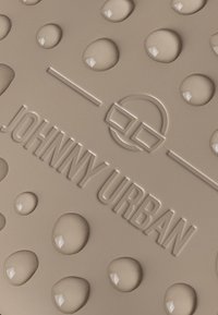 Beige matta med upphöjda droppar och präglad "JOHNNY URBAN"-logotyp, med en slät textur och runda detaljer.