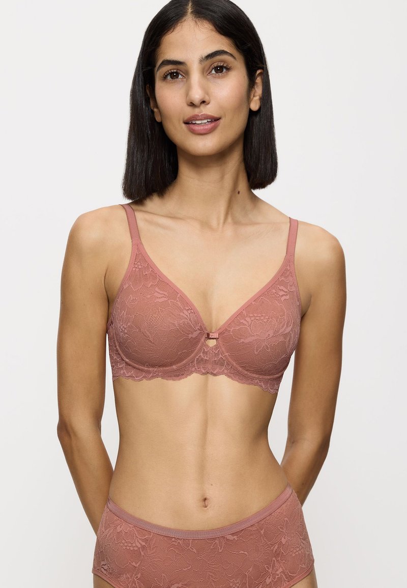 Donna con capelli neri lisci che indossa un completo di pizzo color rosa coordinato, composto da reggiseno e slip, su uno sfondo chiaro e uniforme.