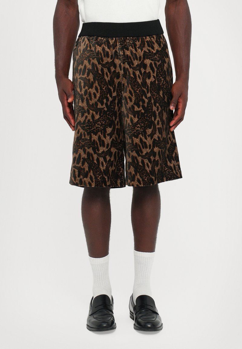 Homme portant un short à motifs marron et noir avec une taille noire, des chaussettes blanches et des mocassins en cuir noirs, se tenant devant un fond uni.