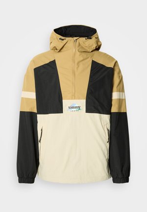 Windbreaker jacket in tan, black, and cream. Features a quarter-zip design, adjustable hood, and side zip pockets. Logo on the chest.