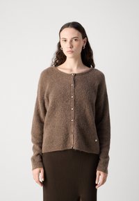 Gilet en tricot marron avec une texture douce et duveteuse, présentant un col rond, des manches longues et une fermeture à cinq boutons. Porté sur une jupe marron côtelée.