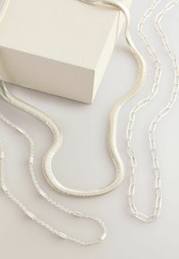 Drei silberne Halsketten, bestehend aus einer flexiblen Schlangenkette, einer Gliederkette und einer kürzeren Kette mit Perlenakzenten, präsentiert auf einer hellen Oberfläche.