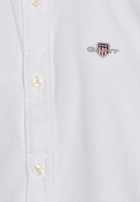 Chemise blanche à boutons, en tissu texturé, avec des boutons de couleur claire et un petit logo brodé en gris, bleu et rouge.