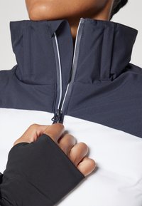 Farbblockierte Jacke in Marineblau und Weiß, mit hohem Kragen, Reißverschluss und strukturiertem Stoff. Die Bündchen haben eine enganliegende Passform.