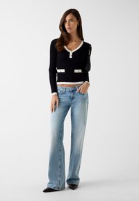 Zwarte geribbelde trui met witte accenten en knopen, gecombineerd met lichtblauwe wijde spijkerbroek. De outfit wordt aangevuld met zwarte, puntige hakken.