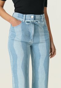 Pantalones de mezclilla de color azul claro con rayas onduladas verticales, cintura alta, cinturón ajustable con lazo y bolsillos laterales. Textura suave con un ligero brillo.