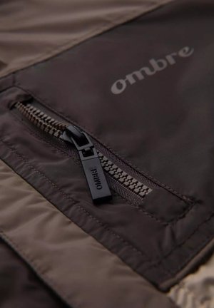Tejido de chaqueta marrón oscuro con un bolsillo con cremallera y un tirador de cremallera negro. El nombre de la marca "ombre" está impreso en un tono más claro.