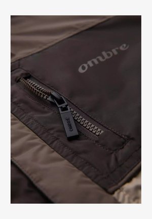 Dark brown jacket fabric featuring a zippered pocket with a black zipper pull. The brand name “ombre” is printed in a lighter shade.