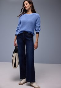 Femme portant un pull bleu en maille, un jean large foncé, des baskets blanches, et tenant un sac cabas rayé beige et noir.