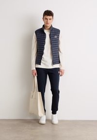 Vest matlasat de culoare bleumarin peste un pulover ușor, asortat cu pantaloni sport negri. Adidași albi și o geantă bej completează ținuta. Design simplu și funcțional.