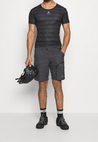 Camisa de manga corta a rayas negras con logo, combinada con pantalones cortos grises que tienen un bolsillo lateral, sosteniendo un casco en la mano, usando zapatos negros con detalles grises.