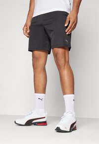 Hombre vestido con pantalones cortos deportivos negros, calcetines blancos de Puma y zapatillas blancas de Puma con acentos en negro y rojo, de pie contra un fondo simple.