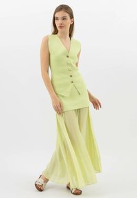 Lichtgroen mouwloos topje met v-hals en knopen aan de voorkant, gecombineerd met een lange, vloeiende, geplooide sheer rok in een vergelijkbare tint. Lichte sandalen.