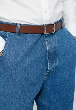 Cintura di pelle marrone con fibbia in metallo argento indossata sopra jeans di denim blu chiaro. I jeans hanno una finitura leggermente scolorita e un design a gamba dritta.