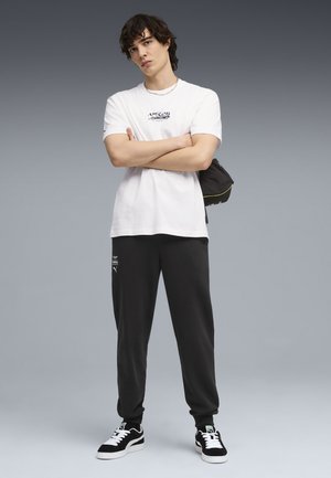 Jonge man die een wit T-shirt met grafische opdruk draagt, zwarte joggingsbroek met logo's, zwart-witte sneakers, met gekruiste armen en een zwarte tas over één schouder.