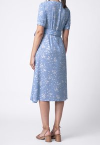 Modré květinové šaty s krátkými rukávy, úzkým pasem a rozšířeným lemem. Výrazné detaily zahrnují jemnou tkaninu a akcent opasku. V kombinaci s béžovými sandály.