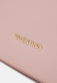 Rozā teksturēta ādas soma ar izteiktu zelta "Valentino" logotipu. Virsma ir ar mīkstu, oļveida apdari.
