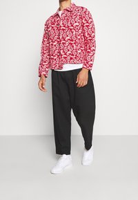 Mann trägt rote Jacke mit Paisley-Muster, weißes Unterhemd, schwarze locker sitzende Hose und weiße Turnschuhe, steht vor weißem Hintergrund.