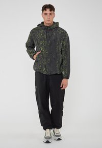 Giacca nera con motivi geometrici verdi, cappuccio e zip frontale. Pantaloni neri in materiale leggero. Il modello è in piedi con le mani nelle tasche.