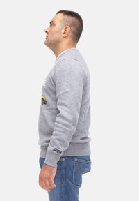 Maglione grigio a maglia con scollo rotondo, polsini a coste e una grafica che presenta un'auto. Indossato con jeans blu.