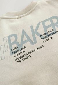 Felpa color crema con grande testo blu che presenta dettagli del marchio e coordinate. Il colletto è texturizzato, offrendo un dettaglio in più.