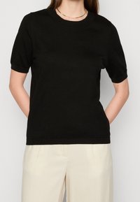 Černé bavlněné pletené tričko s krátkým rukávem a kulatým výstřihem, nošené přes krémové kalhoty. Minimalistický design, bez vzorů.