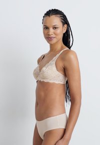 Beige kanten bralette met bloemenpatroon, driehoekige cups en dunne verstelbare bandjes, gecombineerd met naadloze huidskleurige onderbroek. Textuurafwerking.