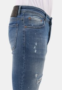 Blauwe denim jeans met versleten kenmerken, met franjeranden en lichte verfspetters. De tailleband toont een zwart etiket.