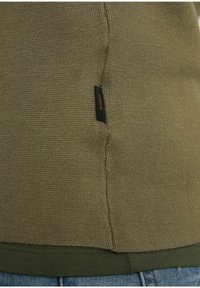 Gros plan de l'ourlet d'un pull tricoté vert olive avec une étiquette en tissu noire portant la mention "CHASIN'" au-dessus d'un sous-pull vert foncé et d'un jean en denim bleu.